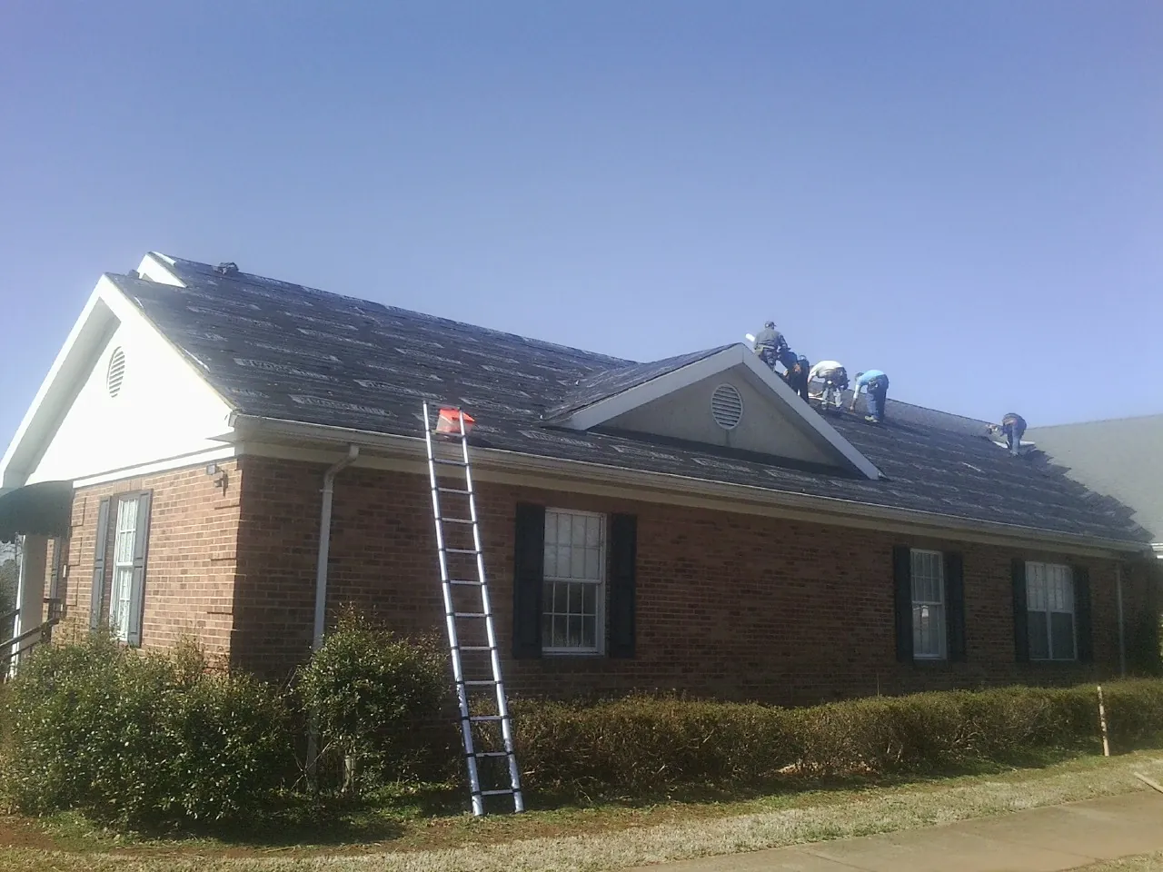 Crew tearing shingles off a brick ranch roof with a ladder set — Metal Roof Repair work by our crew in Hassell