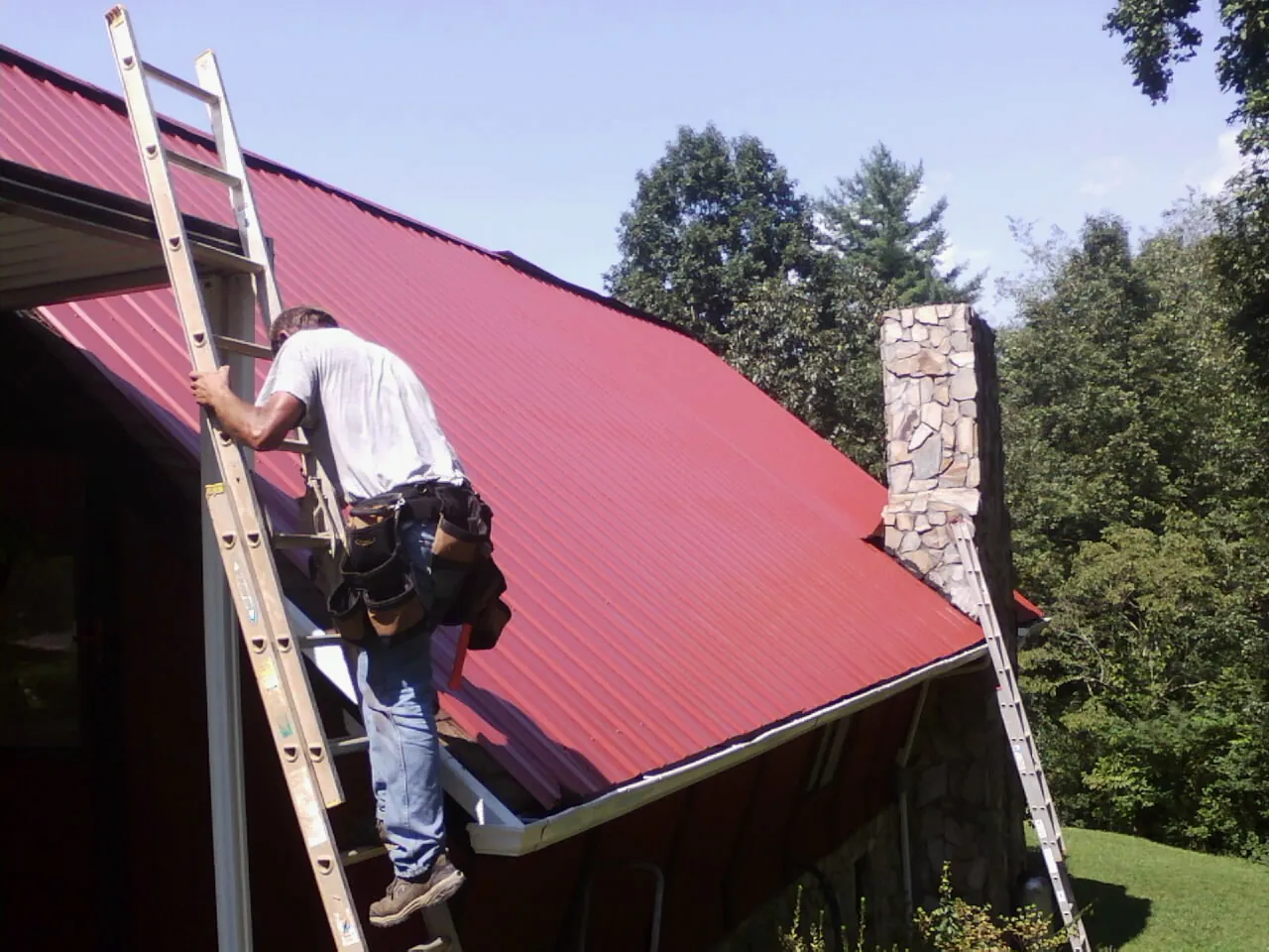 Roofer on a ladder beside a red metal roof and stone chimney — Soffit & Fascia Repair by Roofing Brothers in Hassell